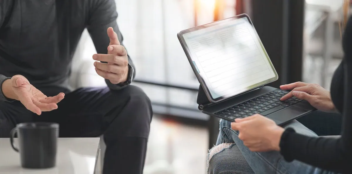 Two professionals in a partner discovery meeting, one gesturing while the other takes notes on a tablet